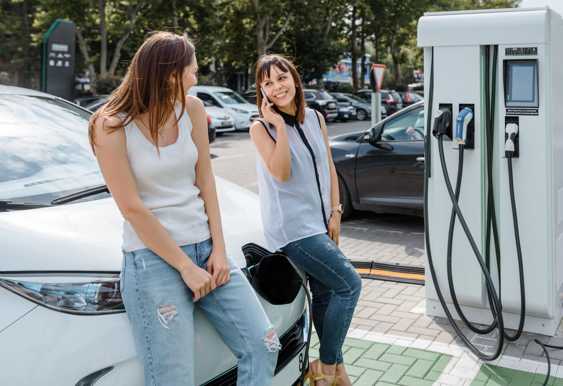 Two happy young beautiful women are talking to each other until their electric car is charging at the charging station situated in the car park.
