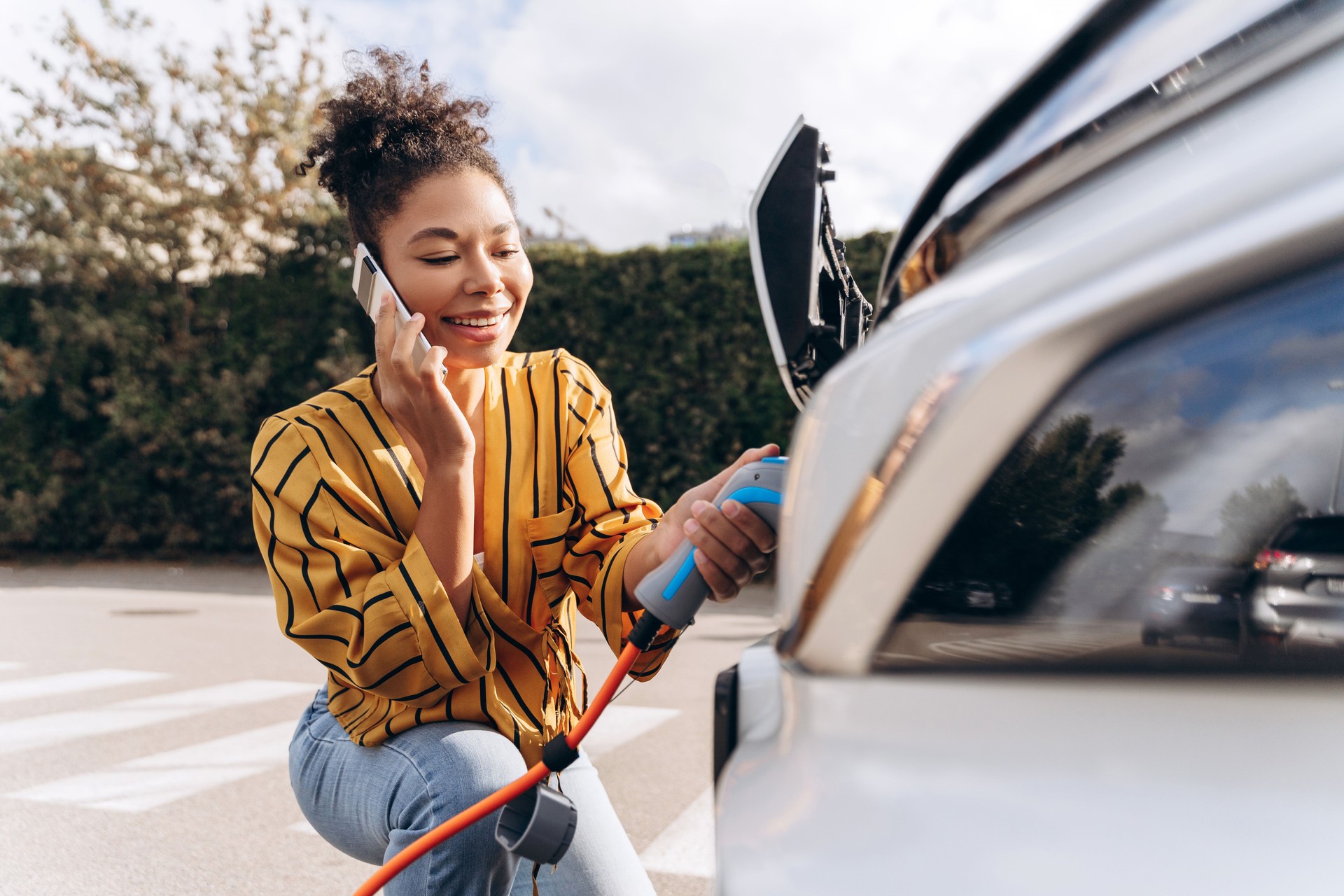 Happy woman inserting EV charger plug into electric car, using mobile phone at charging station